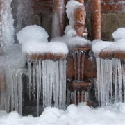 Frozen pipes on an exterior wall of a home, covered in frost and ice, illustrating potential cold weather damage to plumbing.