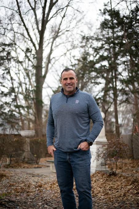 Man in a blue sweater standing outdoors in a wooded area.